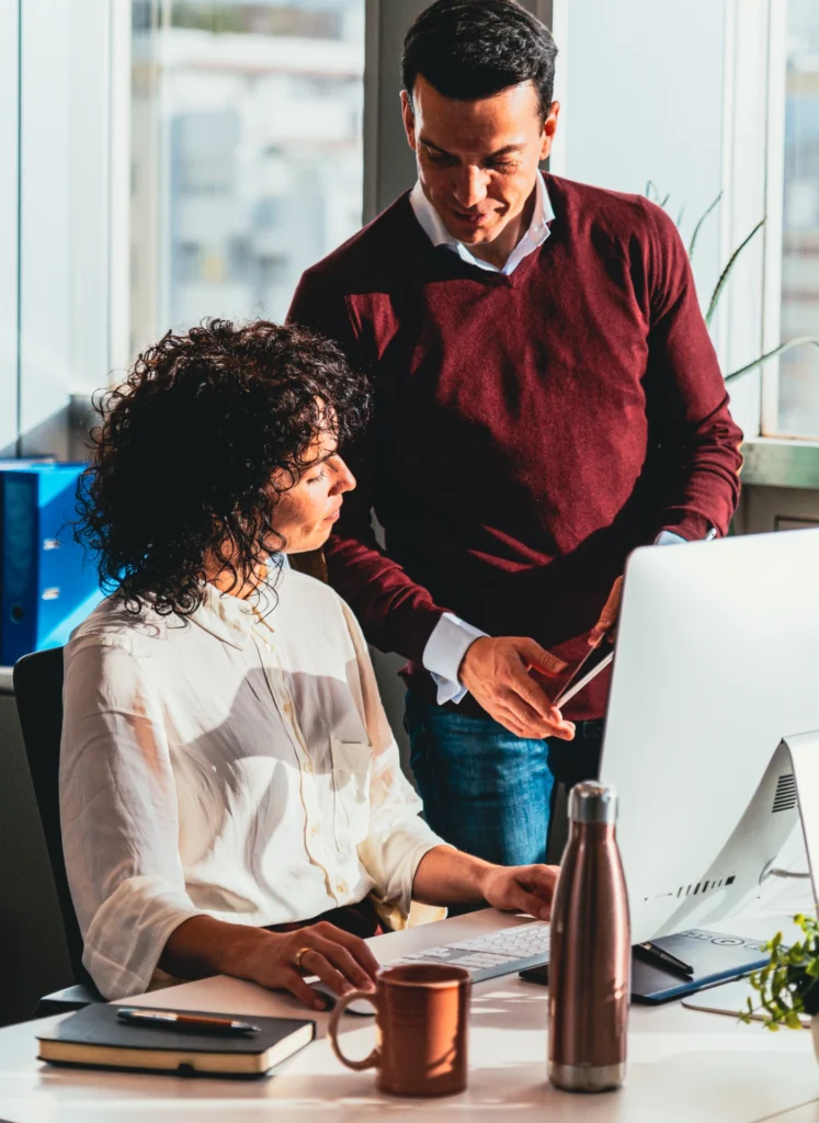 Two colleagues collaborate at a desk by a computer in a bright office.
