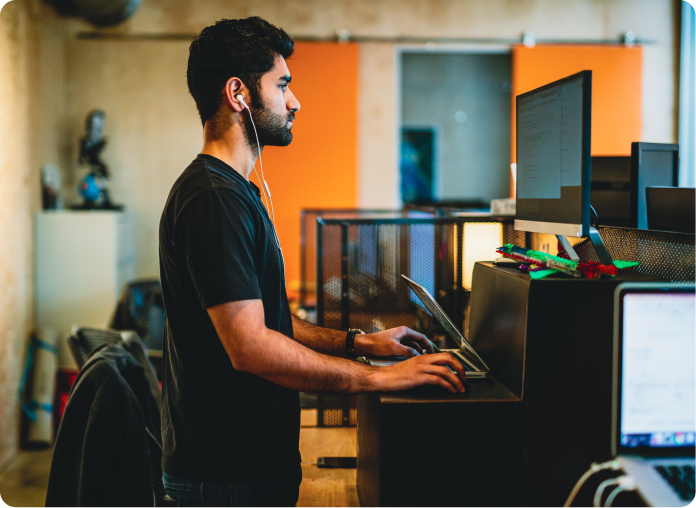 Man wearing earphones stands at a standing desk, typing on a laptop with a large monitor in a modern open office.