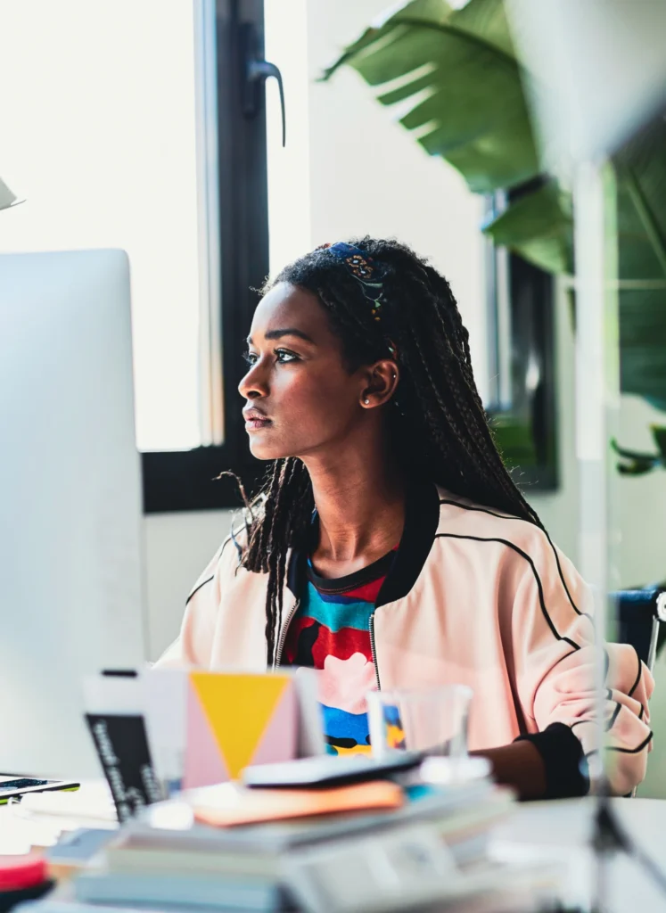 Woman with braided hair sits at a desk in a bright office, looking to the left toward a computer monitor with greenery in the backgrounding plant.