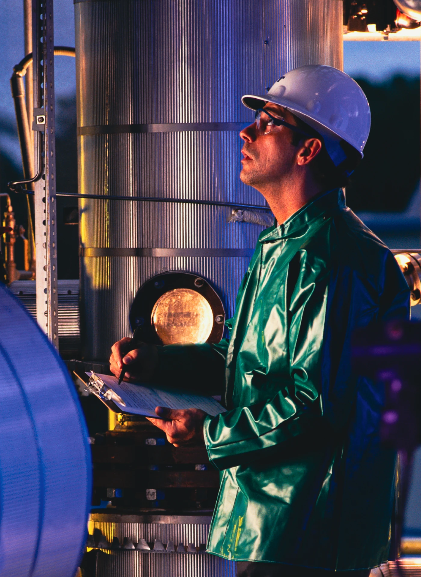 Industrial worker in a white hard hat and green protective suit writes on a clipboard beside large metallic equipment at a facility.