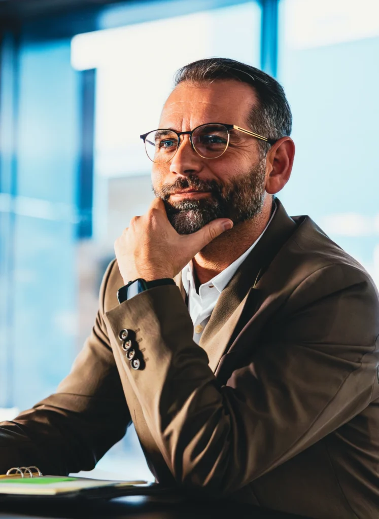 Man with glasses and a beard sits at a desk, chin resting on his hand, looking thoughtful by a bright window.