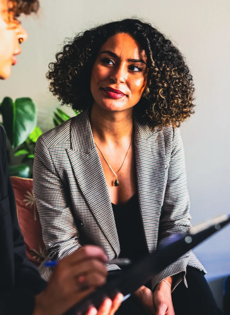 Woman with curly hair in a checkered blazer smiles while talking to a colleague in a bright room.