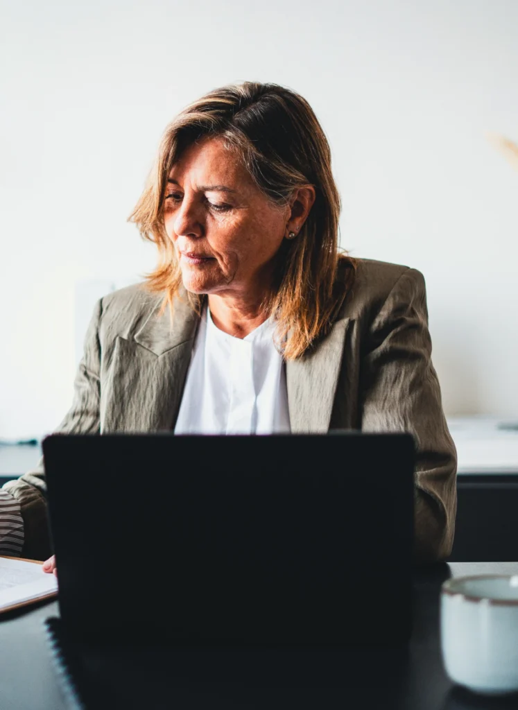 Woman in a blazer sits at a desk, focused on typing on a laptop with papers nearby in a bright office.