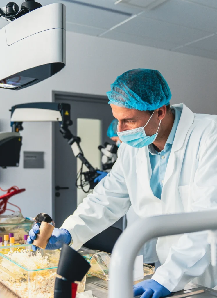 Surgeon in a blue cap and mask performing a sterile lab procedure at a workstation with equipment nearby, in a clinical setting.