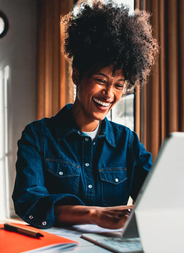 Smiling woman with curly hair working on a laptop at a desk in a denim shirt and orange folders nearby.