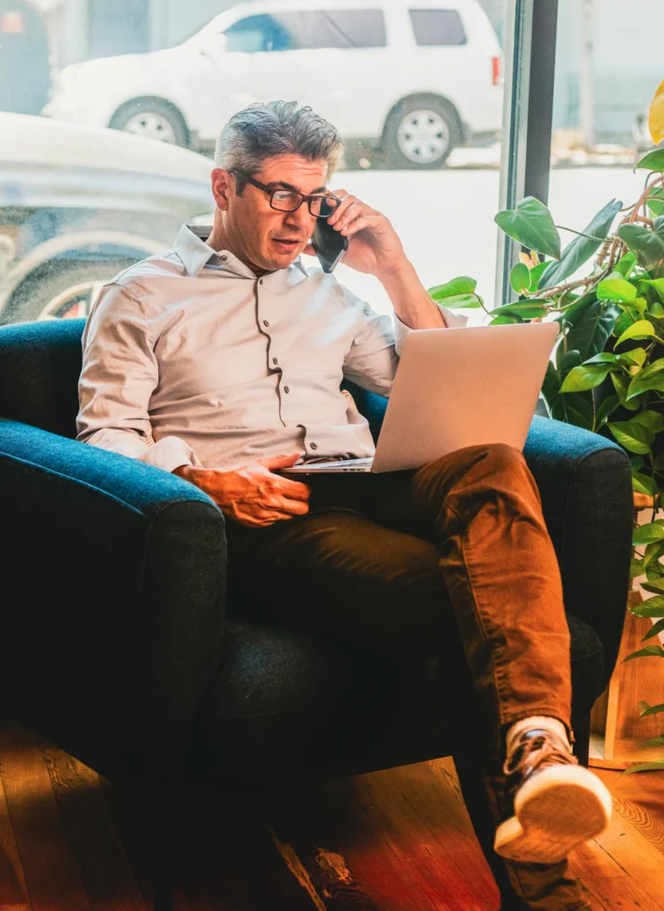 Man in glasses sits on a blue chair, talking on his cellphone with a laptop open on his lap by a window with plants nearby.