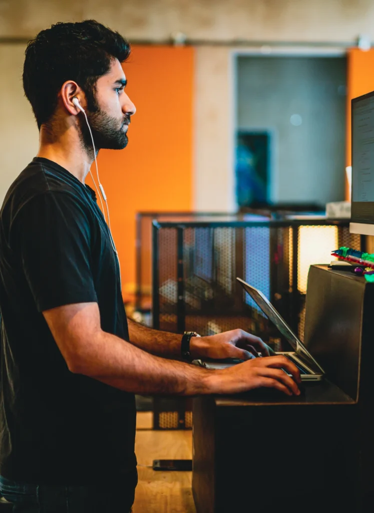 Profile view of a man with earphones typing on a laptop at a workstation in an office.