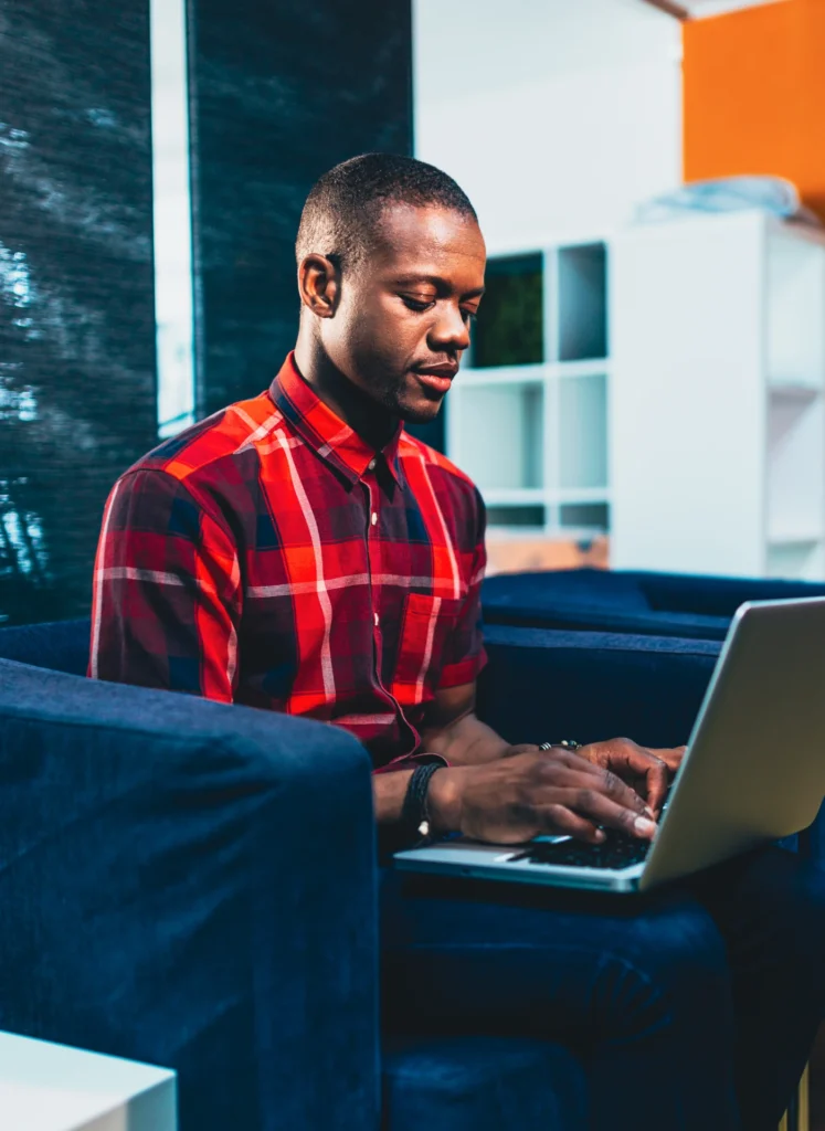Man in a red plaid shirt typing on a laptop while seated on a blue sofa in a modern living room/office space.