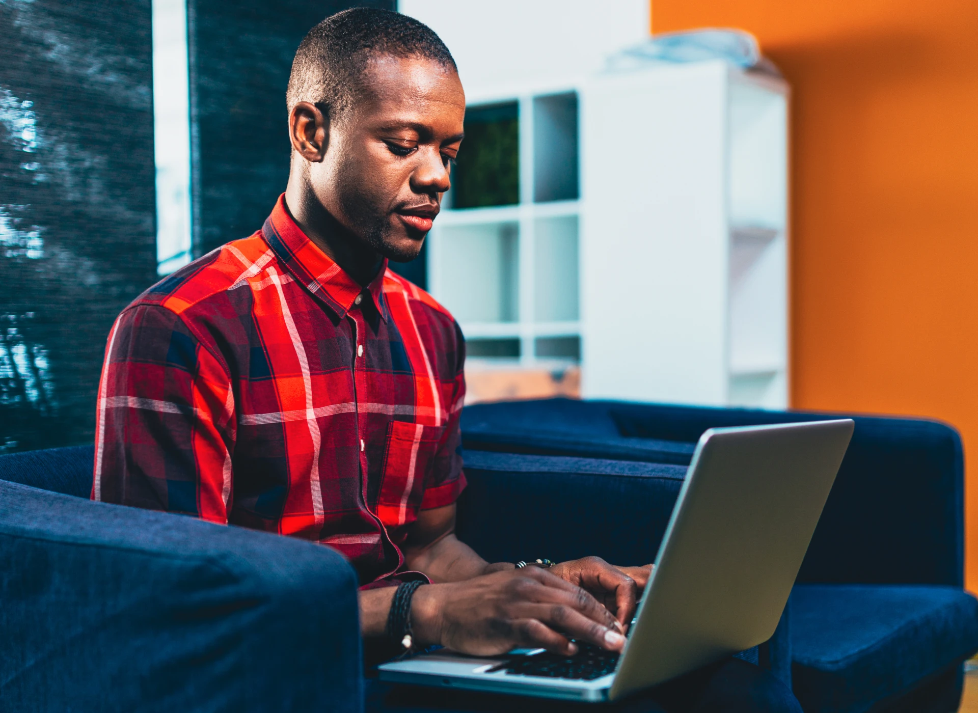 Man in a red plaid shirt typing on a laptop while sitting on a blue sofa.